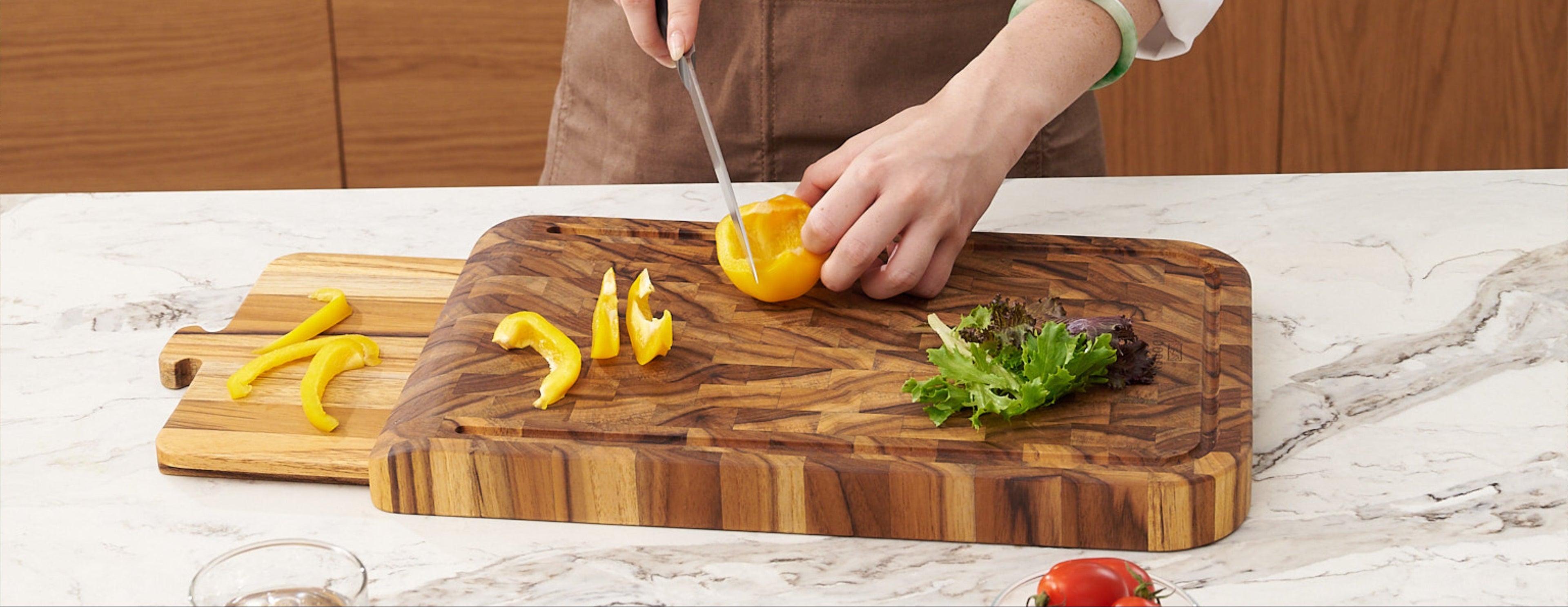 Person cutting a yellow pepper on a Teak wooden cutting board with a marble countertop and wooden cabinets in the background.